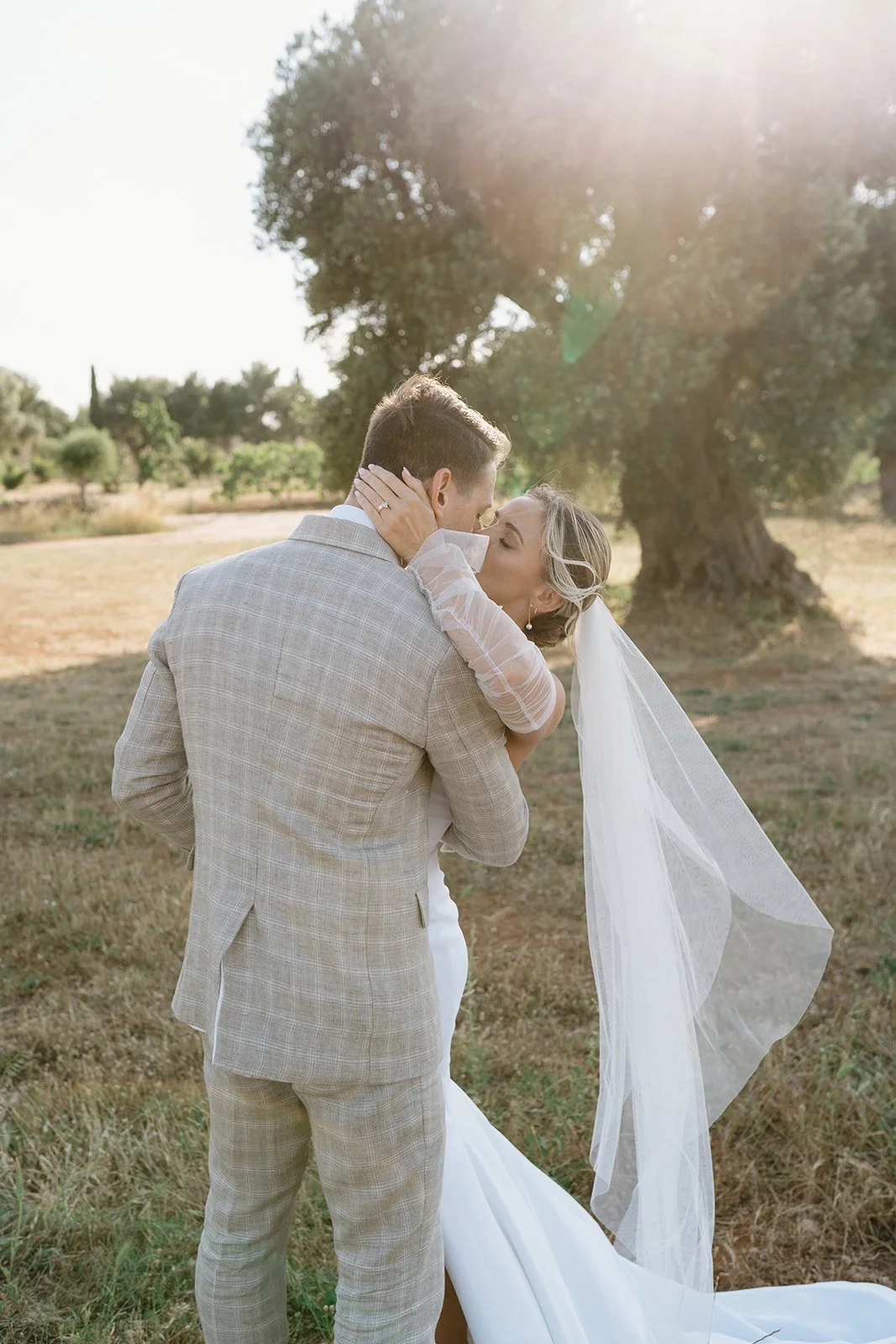 A bride and groom share a romantic embrace outdoors in a sunny, wooded area, with the bride wearing a lace wedding dress and veil, and the groom in a light-colored plaid suit.