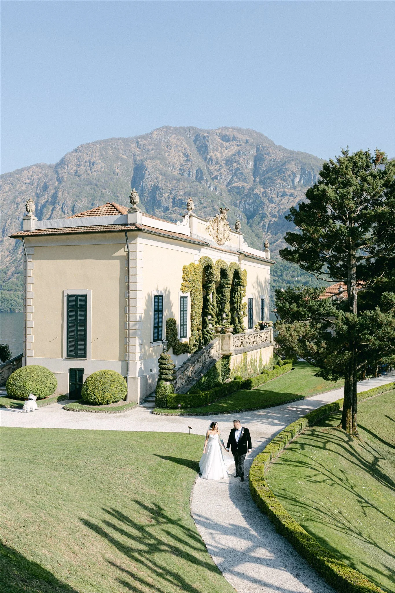 A bride and groom walking hand in hand on a manicured lawn with a historic building and mountain backdrop.