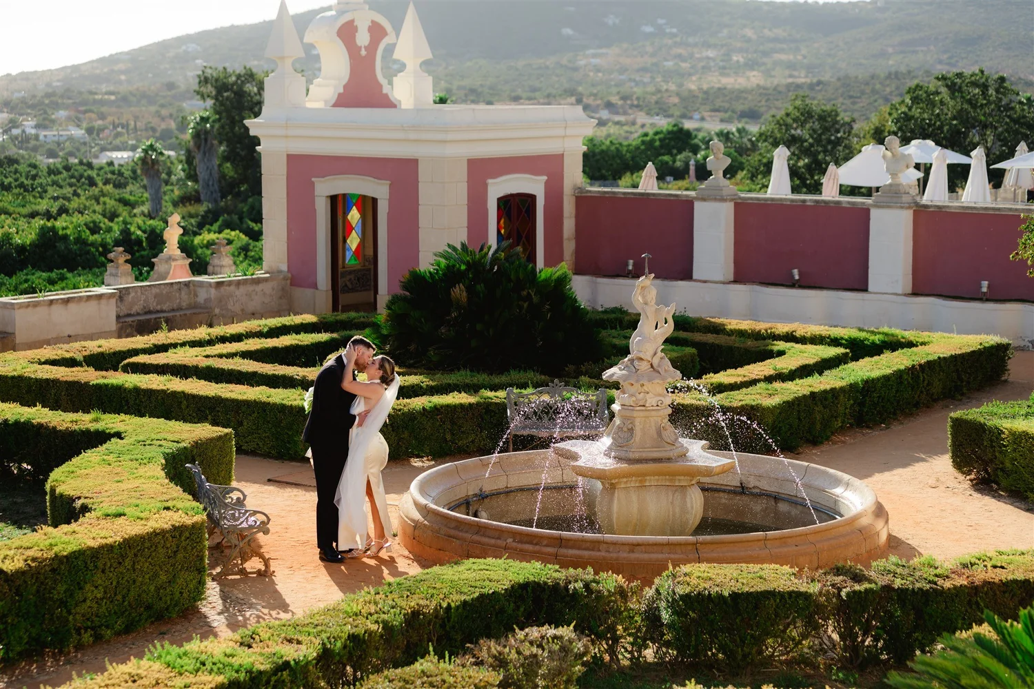 A bride and groom dancing in a formal garden with a fountain and ornate statues, surrounded by lush greenery and historic overlooking mountains.