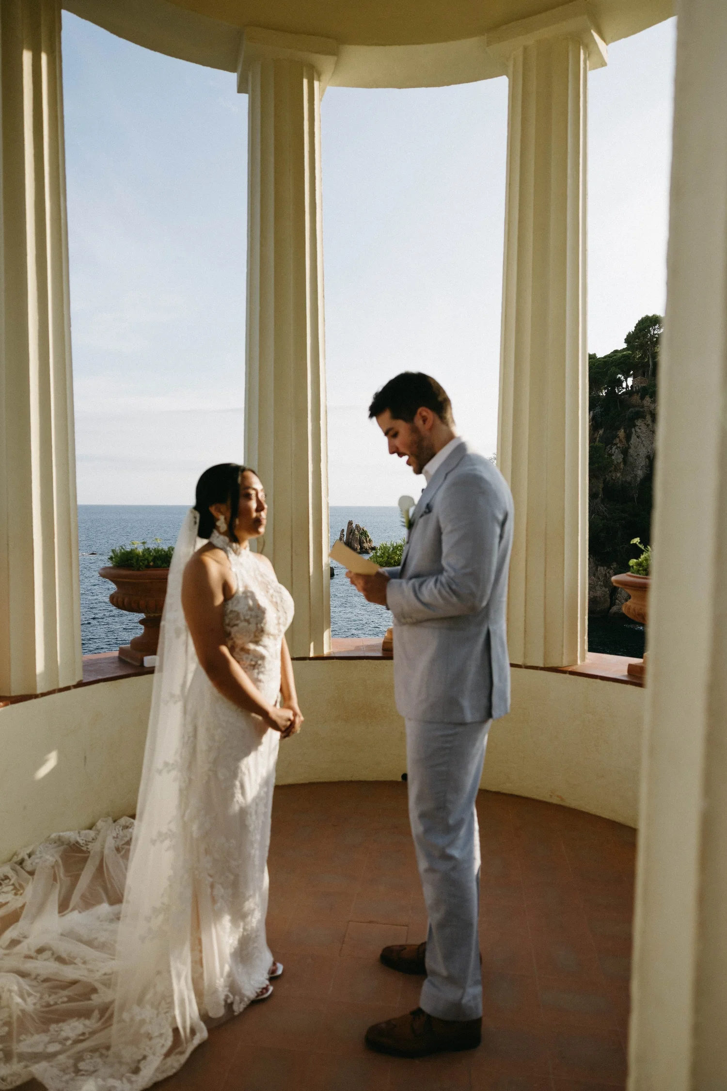 A bride and groom standing face to face during their wedding ceremony on a balcony with an ocean view in the background. The bride is wearing a lace wedding dress with a veil, and the groom is in a light gray suit reading from a paper.