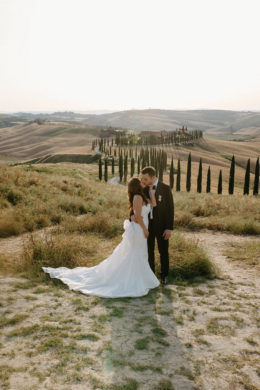 A bride and groom share a kiss in a scenic countryside with rolling hills and a pathway lined with tall cypress trees.