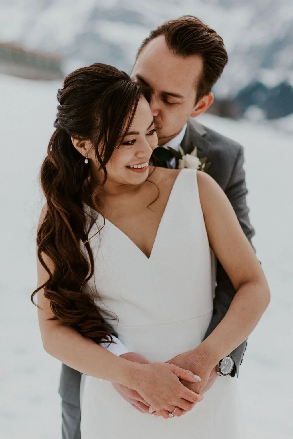 A newlywed couple in wedding attire embracing outdoors on a snowy day, with the groom kissing the bride's temple.