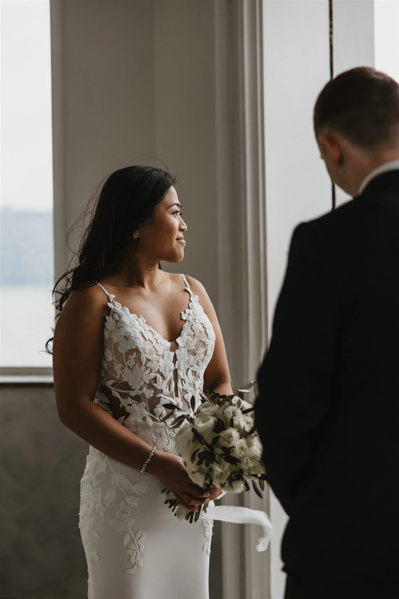 A bride in a white wedding gown with floral lace details looking at a groom in a black suit. The bride is holding a bouquet of white flowers and greenery. They are indoors near a window with natural light.