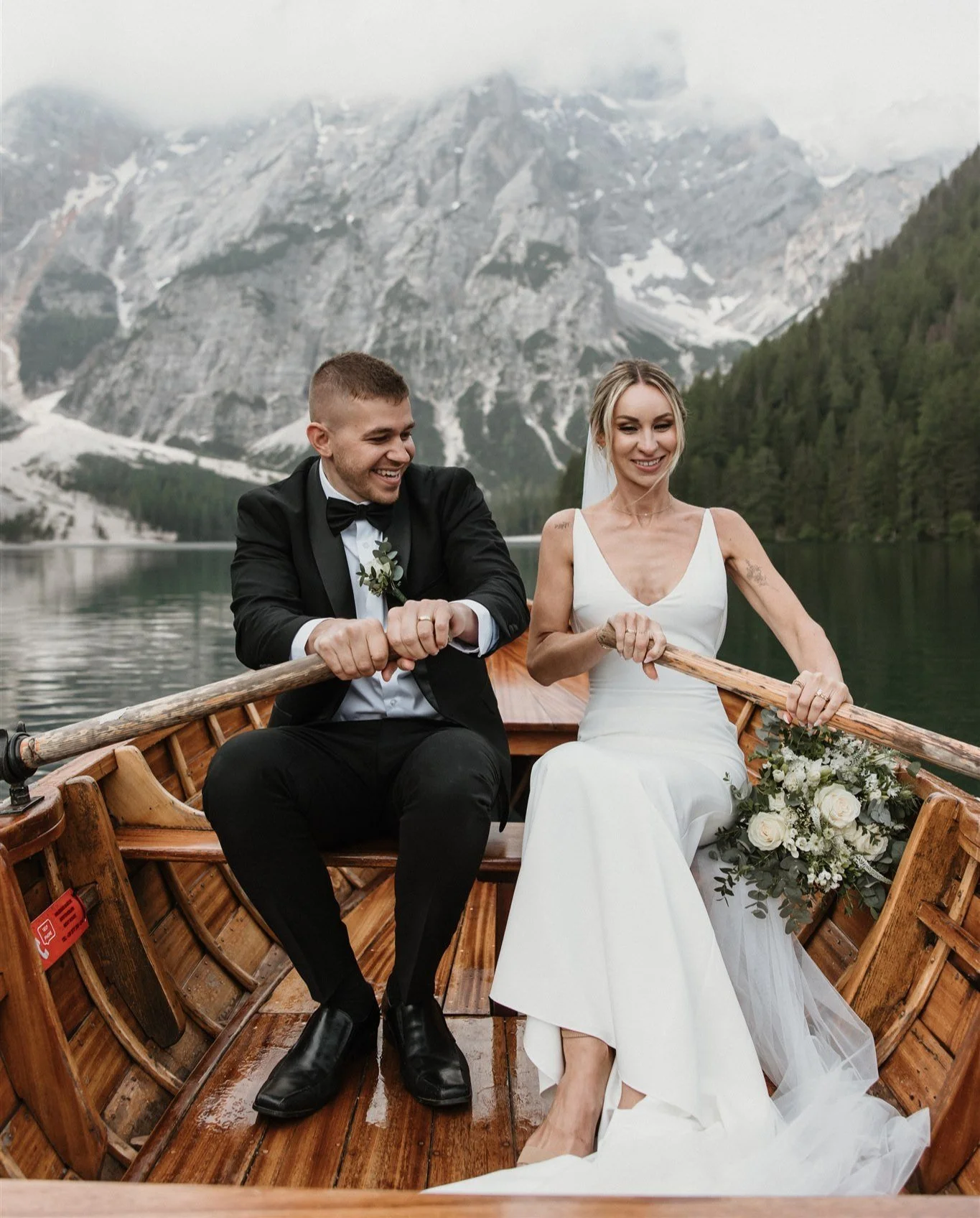 A bride and groom sitting in a wooden boat on a lake surrounded by mountains and trees, with the bride holding a bouquet of flowers.