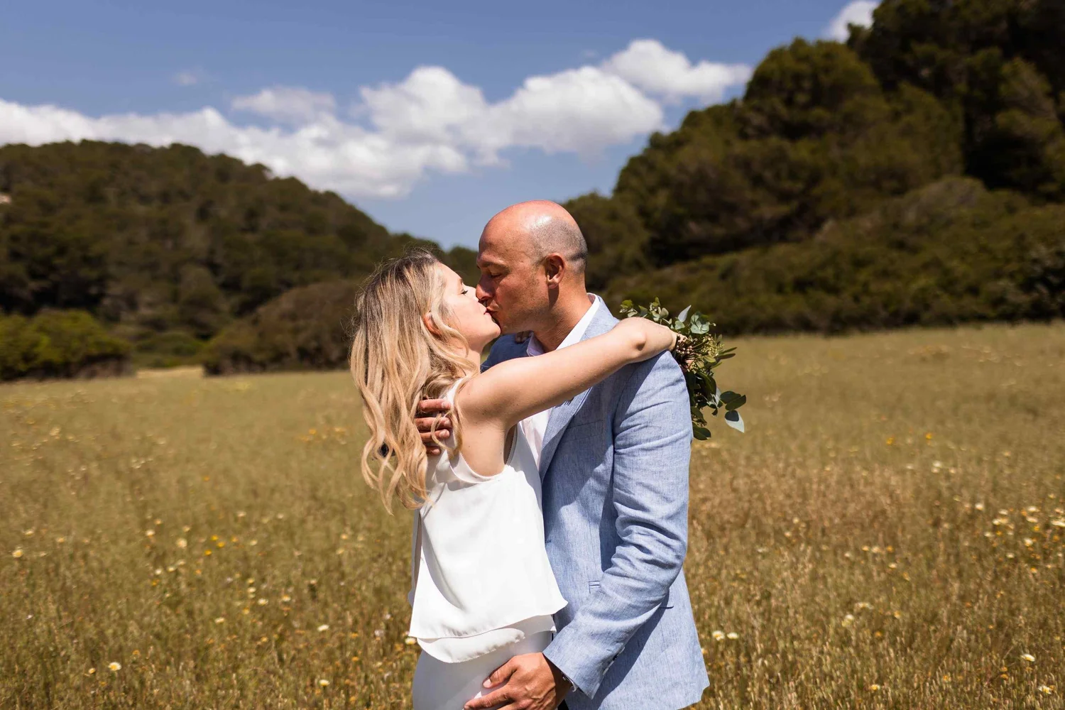 A couple is kissing in a grassy field with hills in the background under a partly cloudy sky.