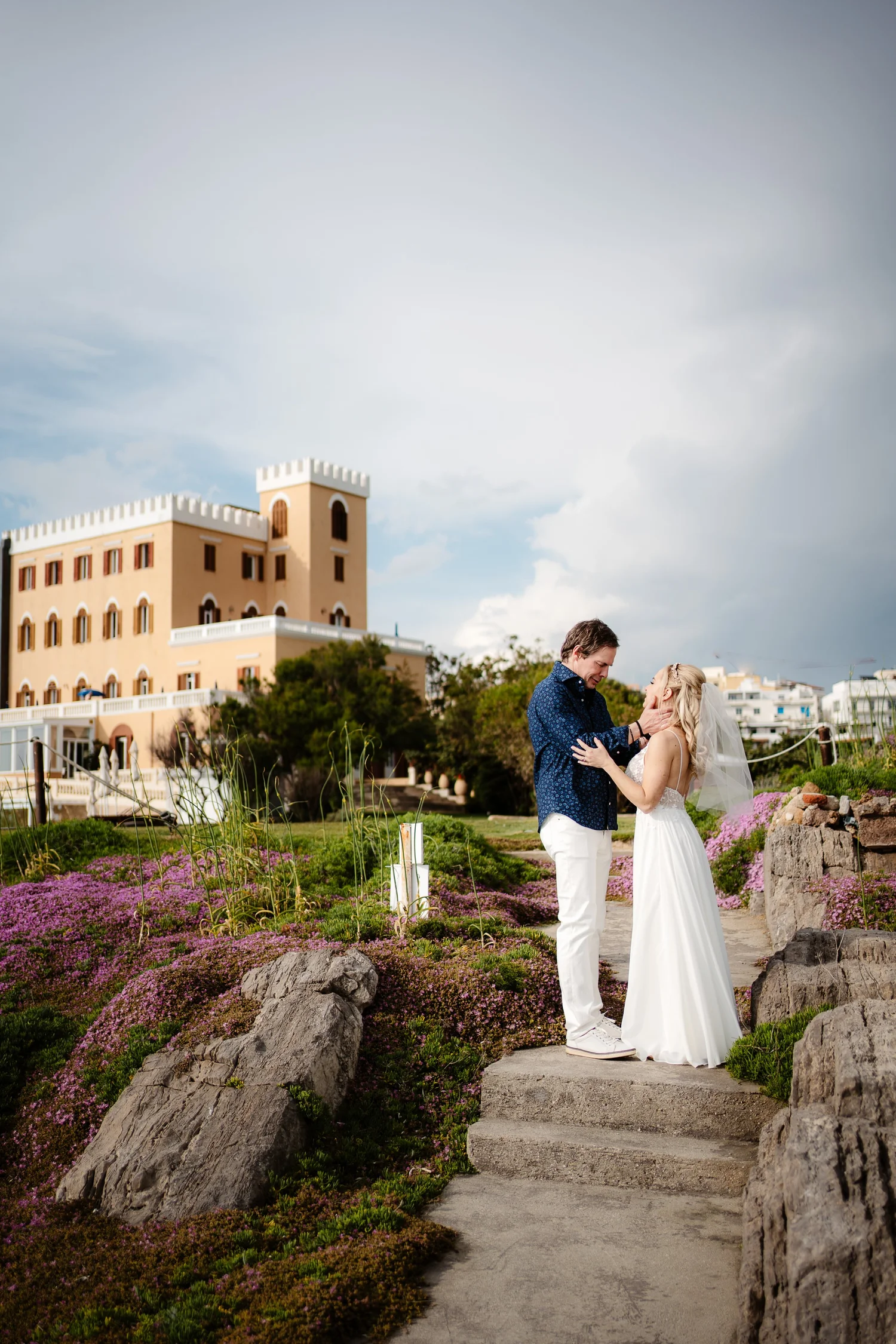 A wedding scene with a bride and groom standing on a stone pathway surrounded by purple flowers and rocks. The groom is holding the bride's face and they are smiling at each other. There is a large beige building with a tower and a blue sky with clou