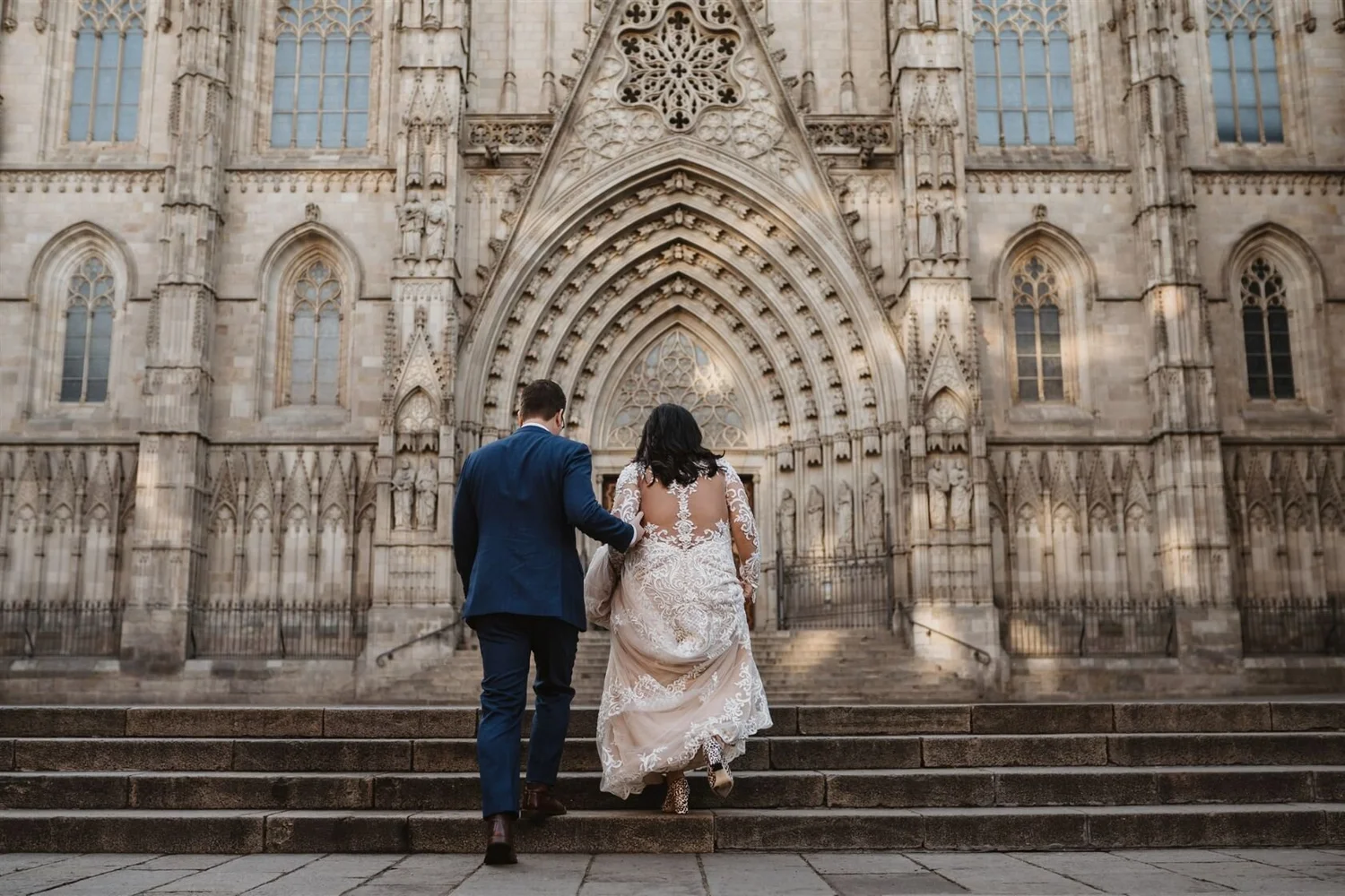 A couple dressed in wedding attire walking up stone steps toward a grand Gothic cathedral, with intricate architectural details and stained glass windows.