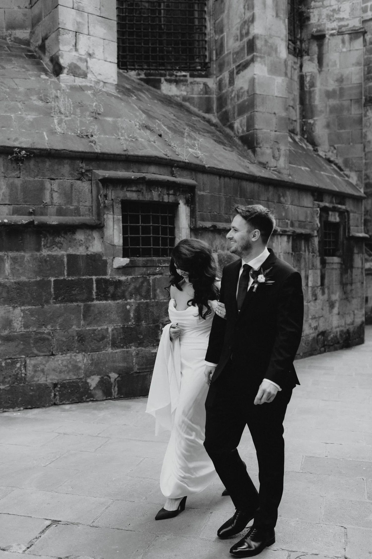 A black and white photo of a bride and groom walking outdoors near a historic brick building. The bride is wearing a long white dress and heels, and the groom is in a suit with a boutonniere. They are smiling and holding hands, enjoying a joyful moment.