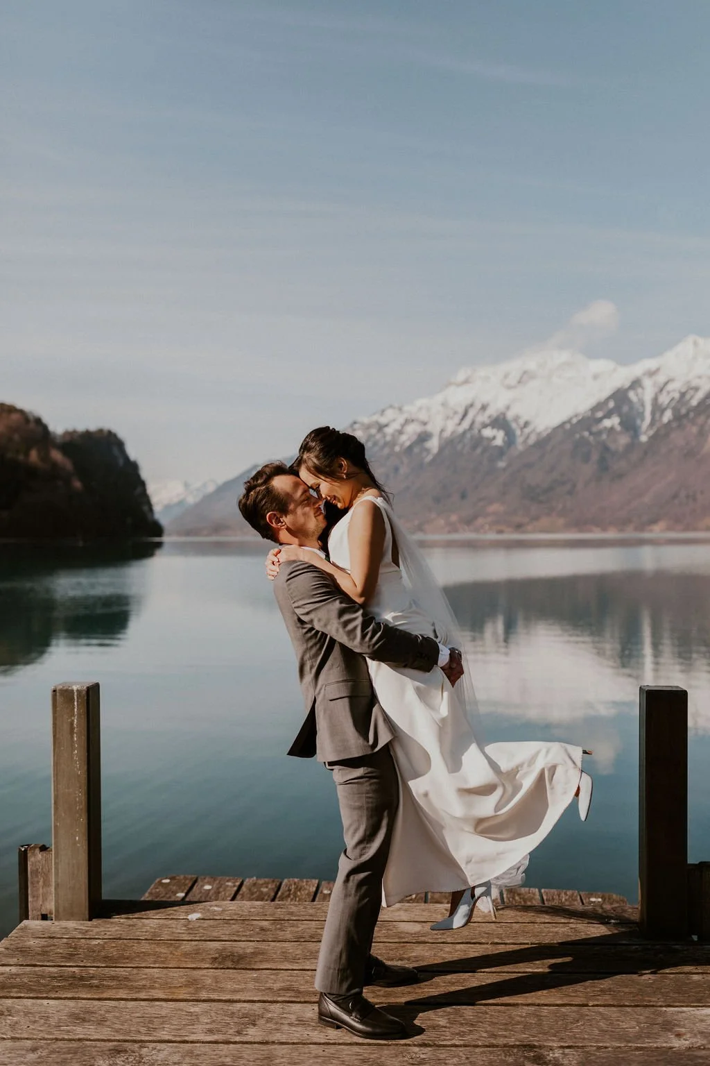 A groom lifting a bride in his arms on a wooden dock by a calm lake with mountain ranges in the background, snow-capped peaks under a blue sky.