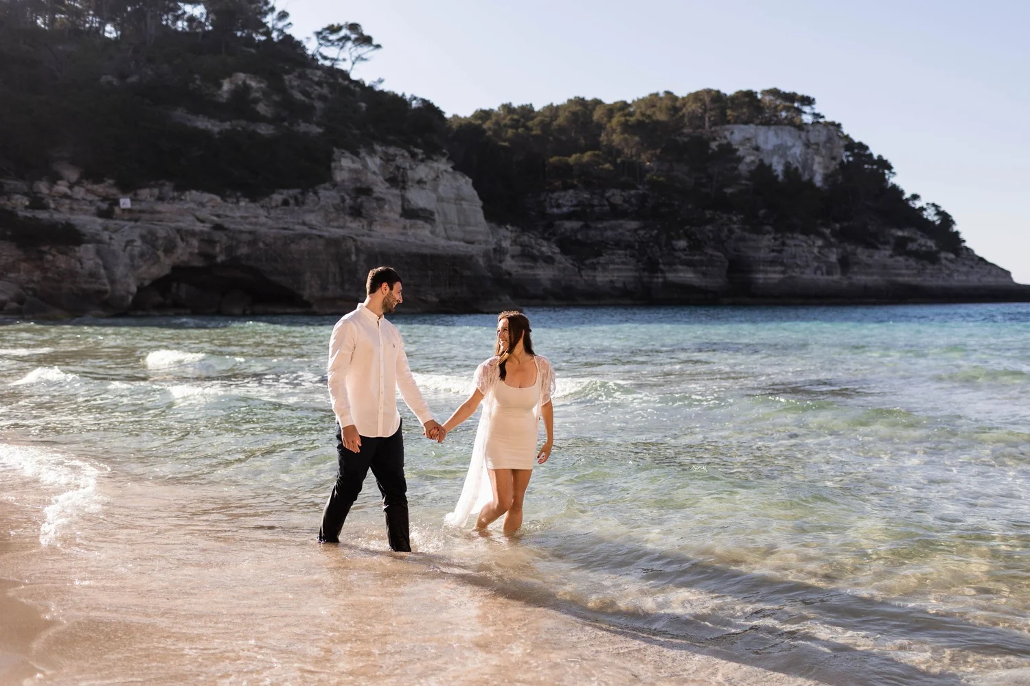 A couple holding hands and walking in shallow ocean water, with a large rocky cliff and trees in the background.