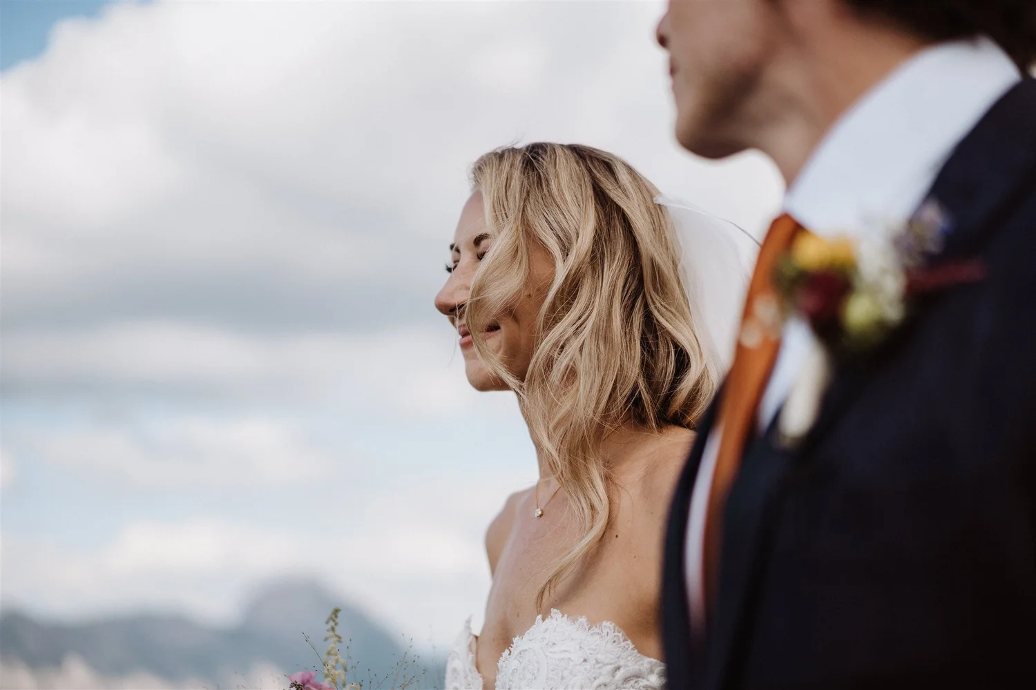Close-up of a bride and groom during a wedding ceremony outdoors, with cloudy sky in the background. The bride has blonde, wavy hair and is smiling, wearing a lace wedding dress. The groom is dressed in a dark suit with a white shirt and a colorful b