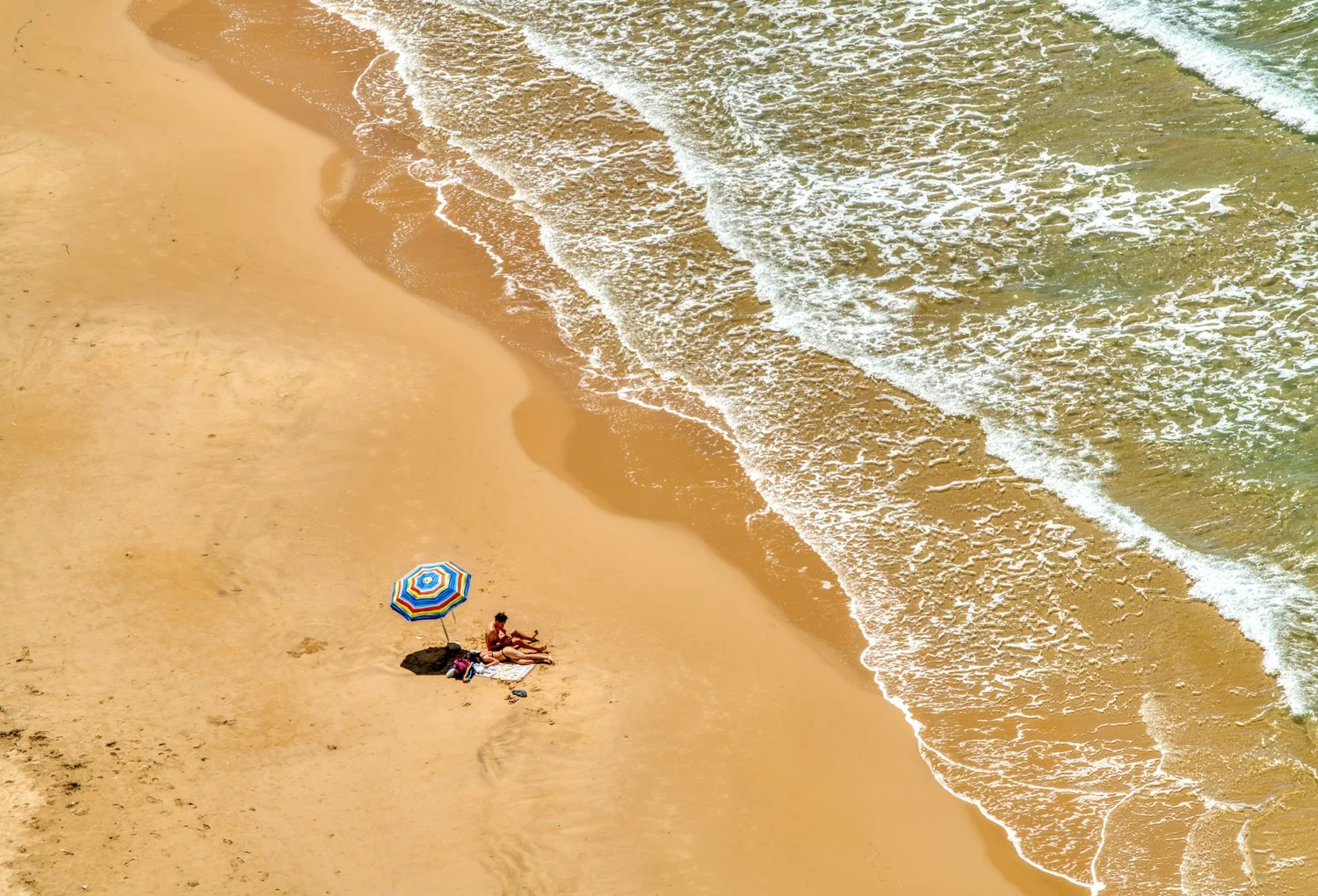 A beach scene from above showing a sandy shore, gentle waves, and a colorful striped umbrella with two people sitting nearby.