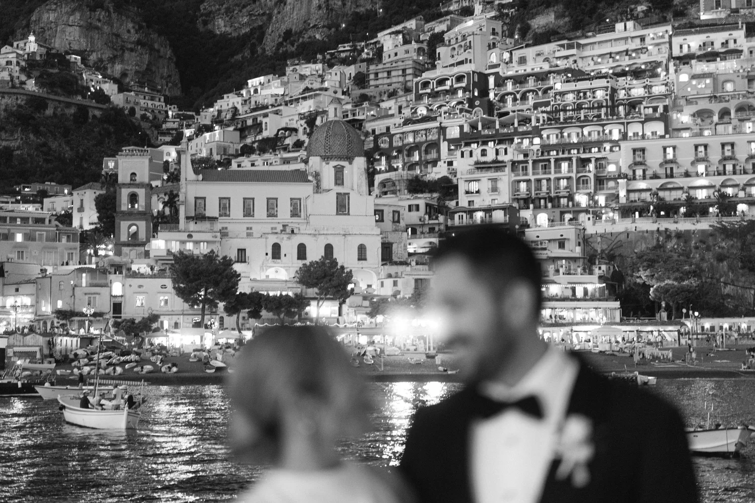 A blurred figure in the foreground with a marina and hillside town in the background at night. The scene includes boats on water and buildings with lights on the hillside.