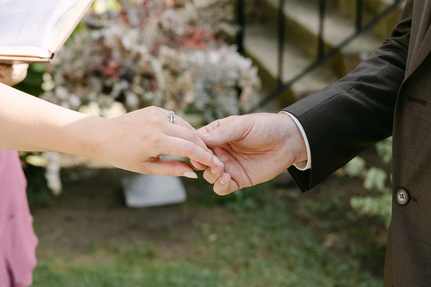Close-up of a couple holding hands during a wedding ceremony, with the bride's hand showing an engagement ring and wedding band, and the groom wearing a black suit.