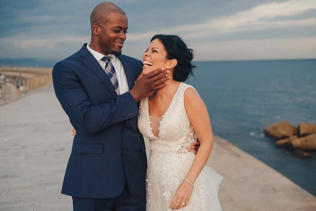 A newlywed couple smiling and laughing by the ocean on a cloudy day, with the groom gently touching the bride's face.