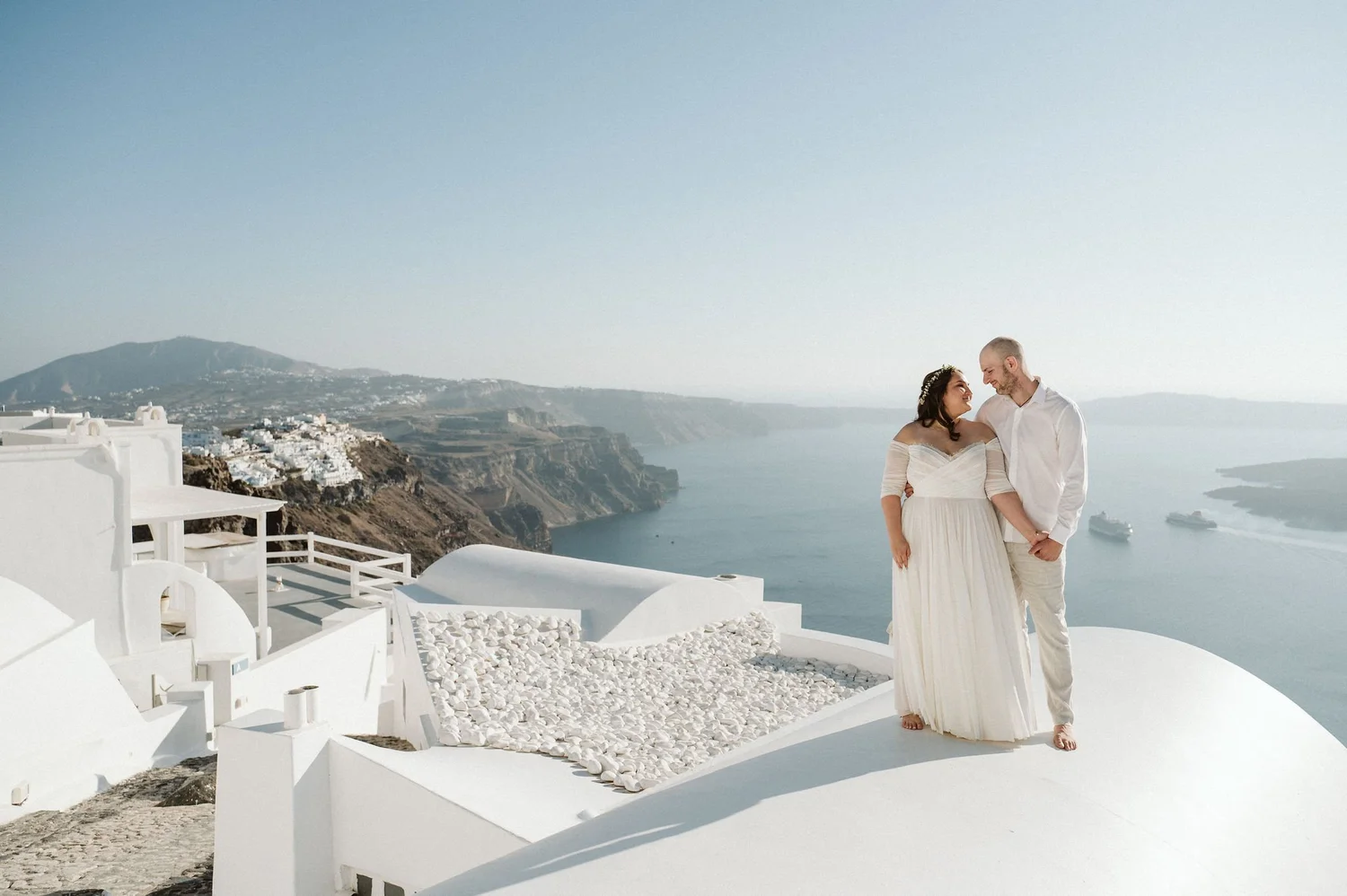 A couple in wedding attire standing together on a white terrace overlooking the ocean, with a scenic coastal landscape and a cruise ship in the water.