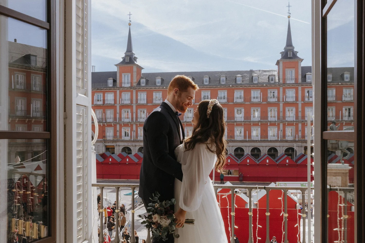 A groom and bride holding hands and smiling at each other on a balcony during their wedding, with a cityscape background of old buildings with red facades and spires
