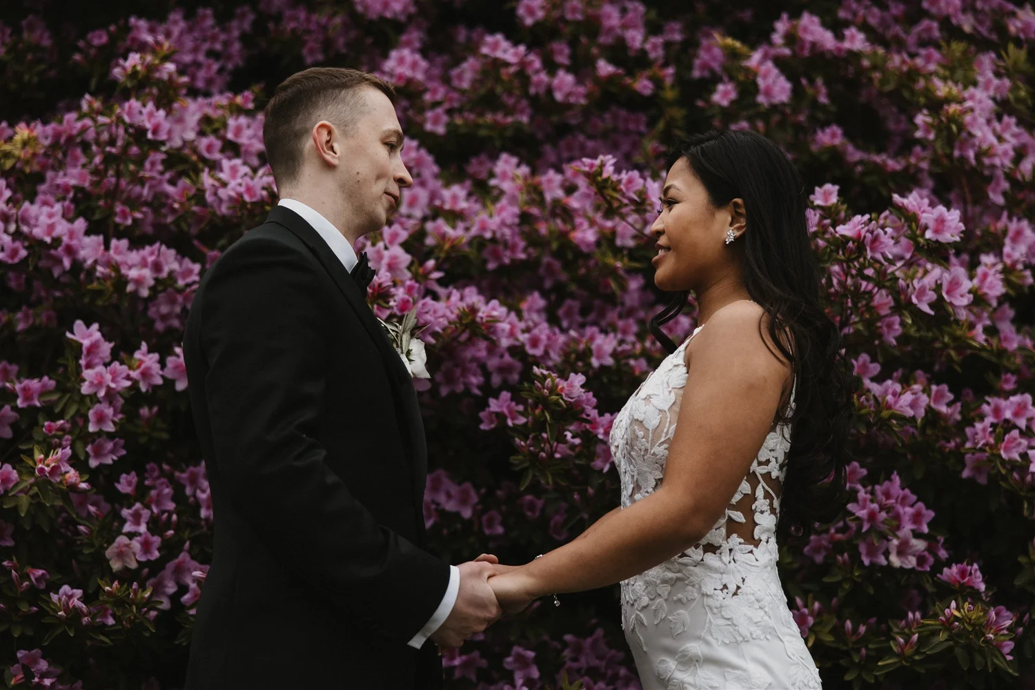 A bride and groom standing face to face, holding hands, with a background of pink azalea flowers.