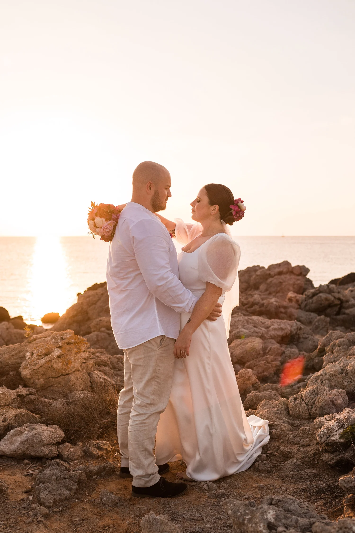 A couple is standing on a rocky beach at sunset, facing each other lovingly. The man has a shaved head and beard, wearing a white shirt and light-colored pants. The woman has dark hair with pink flowers in it, wearing a white dress. The woman is holding a bouquet of pink and orange flowers behind the man's shoulder. The ocean and a setting sun are visible in the background.