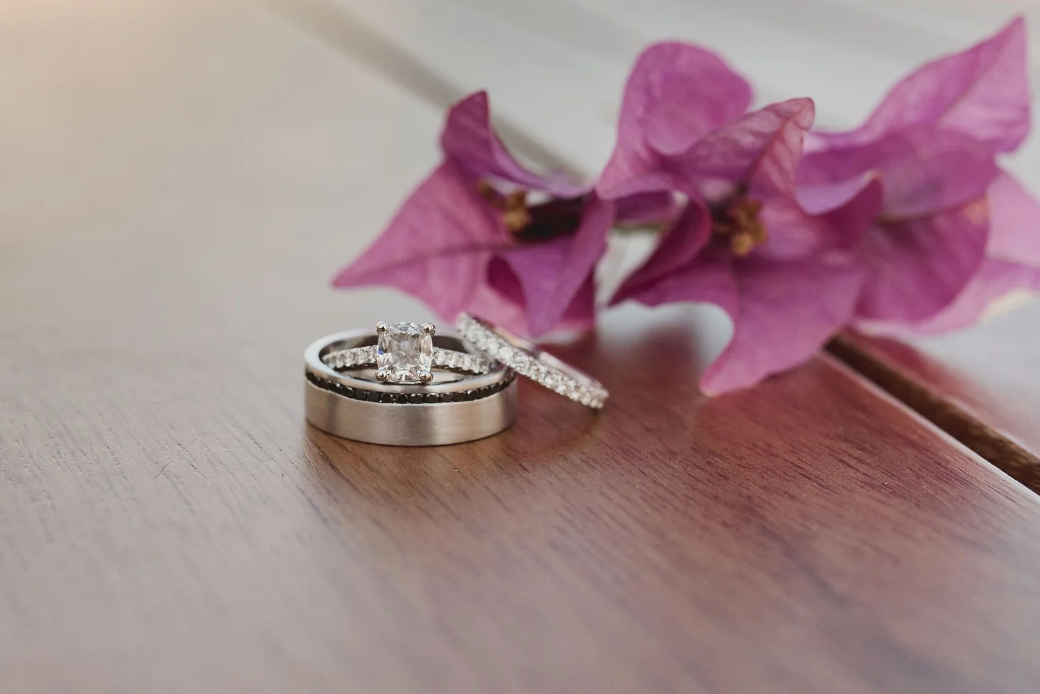 Close-up of a diamond engagement ring on a wooden surface with a pink flower in the background.