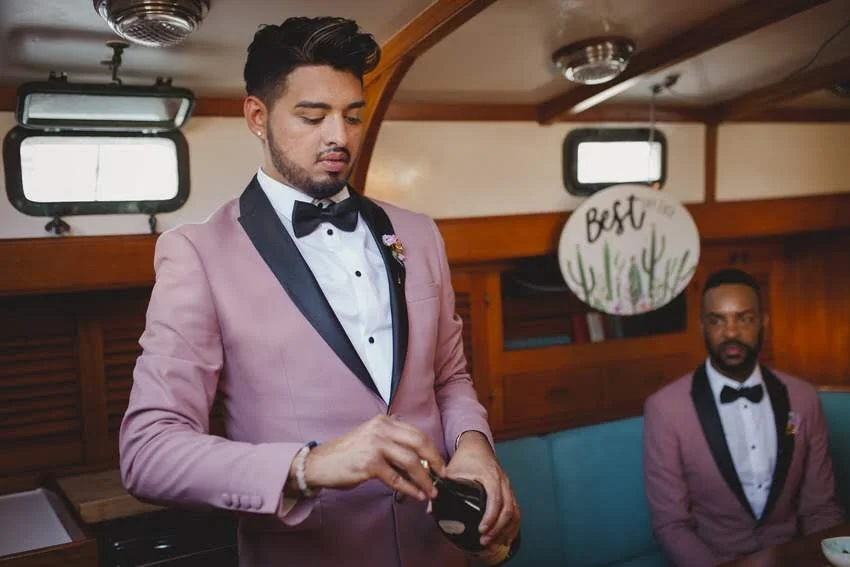 Two men in pink tuxedos with black lapels and bow ties, sitting inside a boat with wood paneling. One man is standing, looking at his phone, while the other is sitting and looking ahead. A round sign that says 'Best' with cactus illustrations is hang