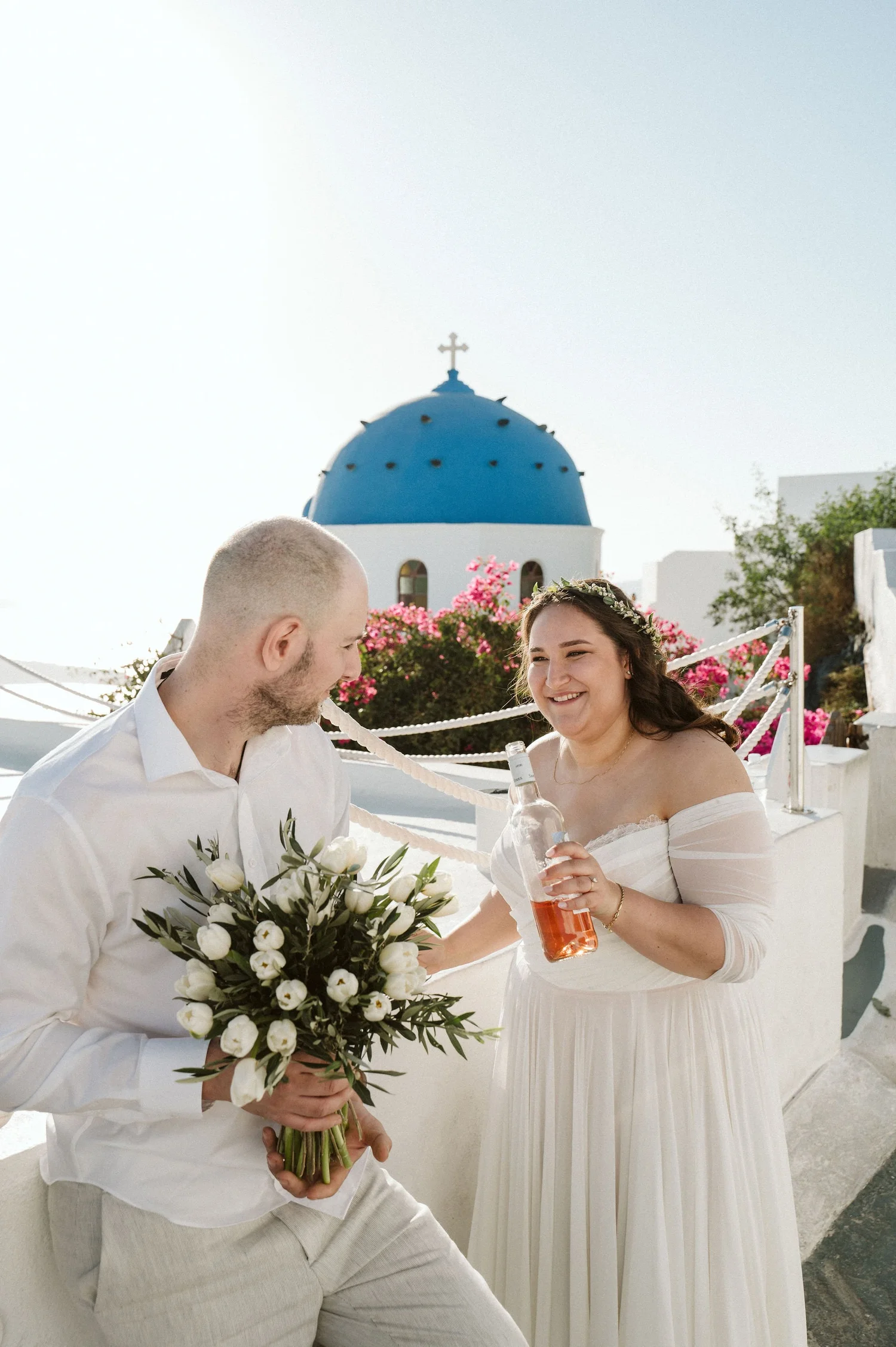 A bride in a white off-shoulder wedding dress and a floral headband holding a drink, smiling at a groom in a white shirt and beige pants, who is holding a bouquet of white roses and greenery; they're outdoors with pink flowers and a blue-domed white