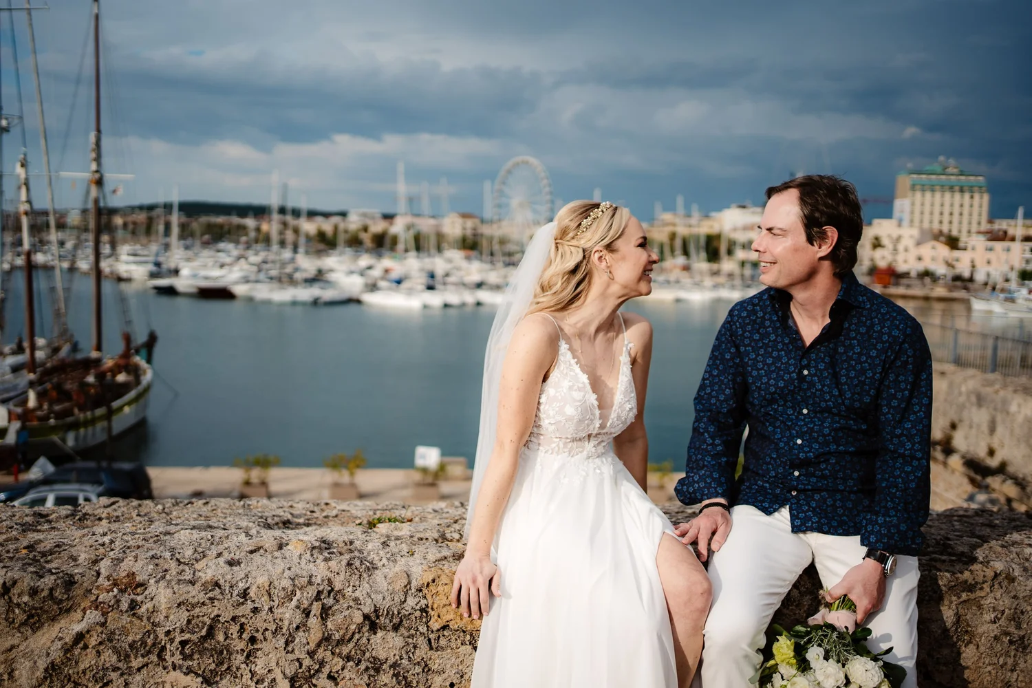 A bride and groom sit on a stone wall near a harbor with boats. The bride wears a white lace wedding dress and a headband, while the groom wears a dark blue shirt with a pattern and white pants. They are smiling and looking at each other, with a city