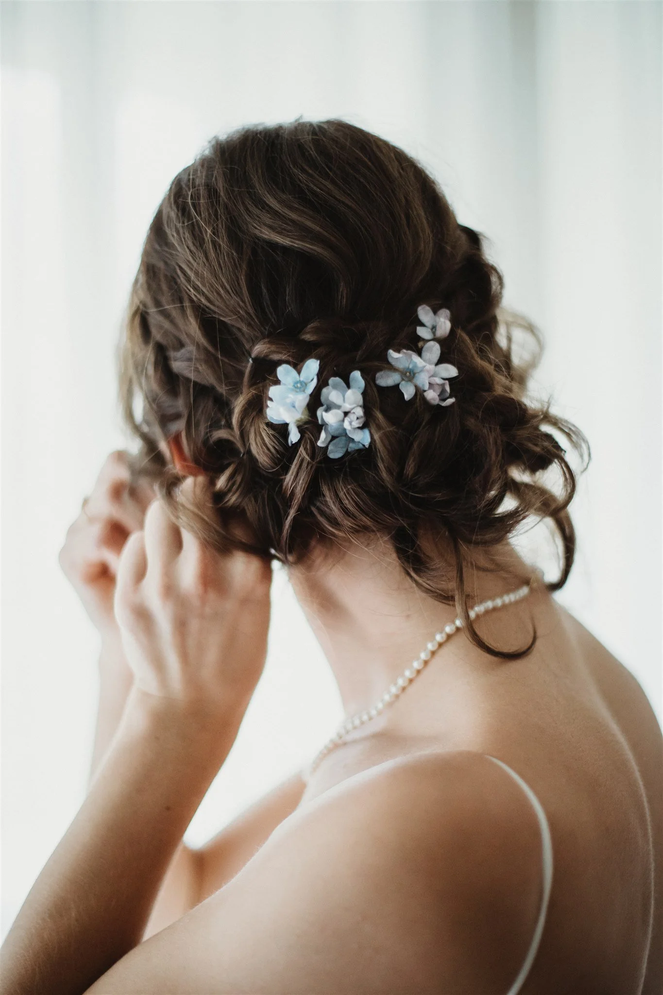Back view of a woman with brown curly hair decorated with blue and white flowers, wearing a pearl necklace and a white spaghetti strap top.