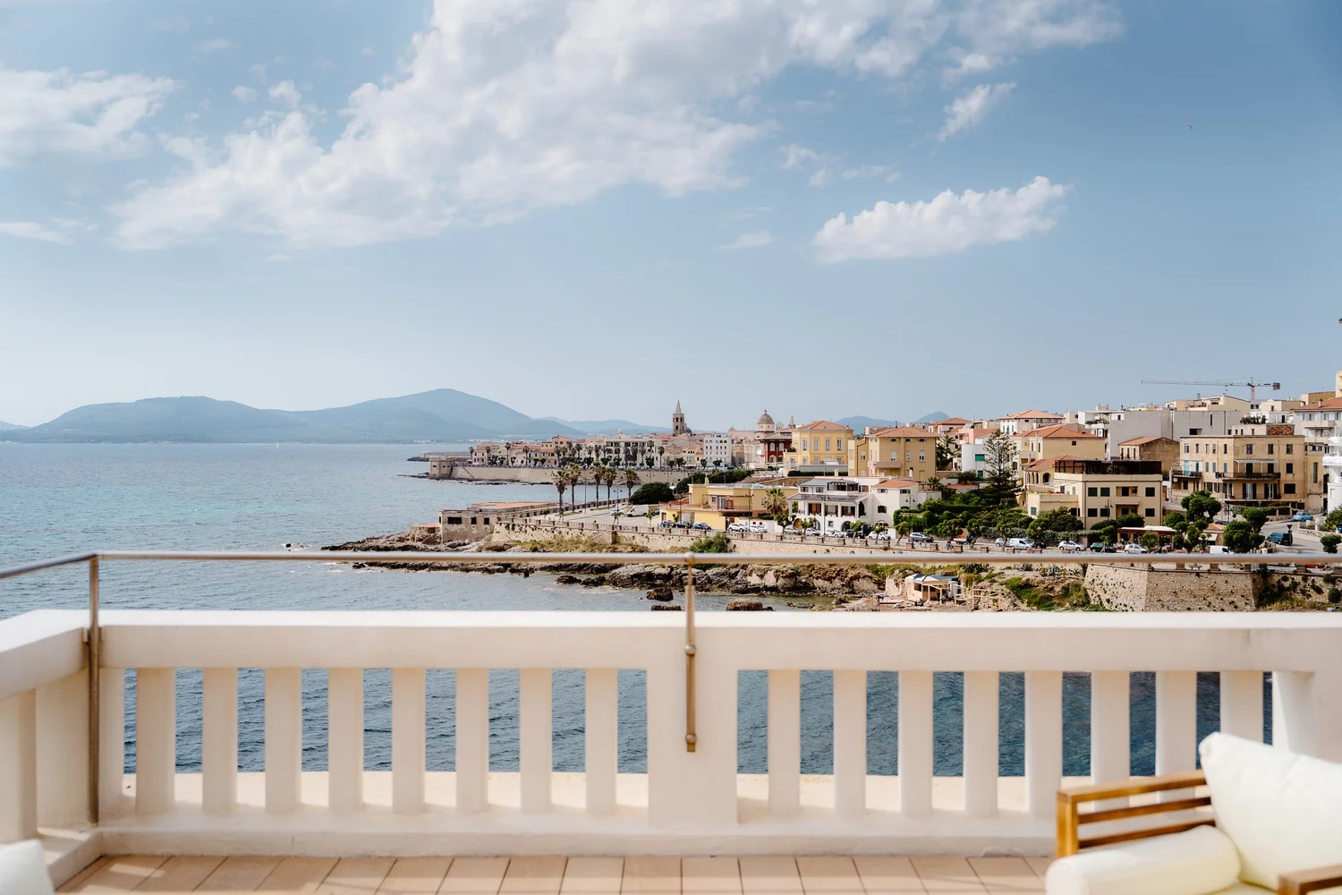 A view of a coastal town with colorful buildings, a church with a tall steeple, and mountains in the background, seen from a balcony with a white railing.