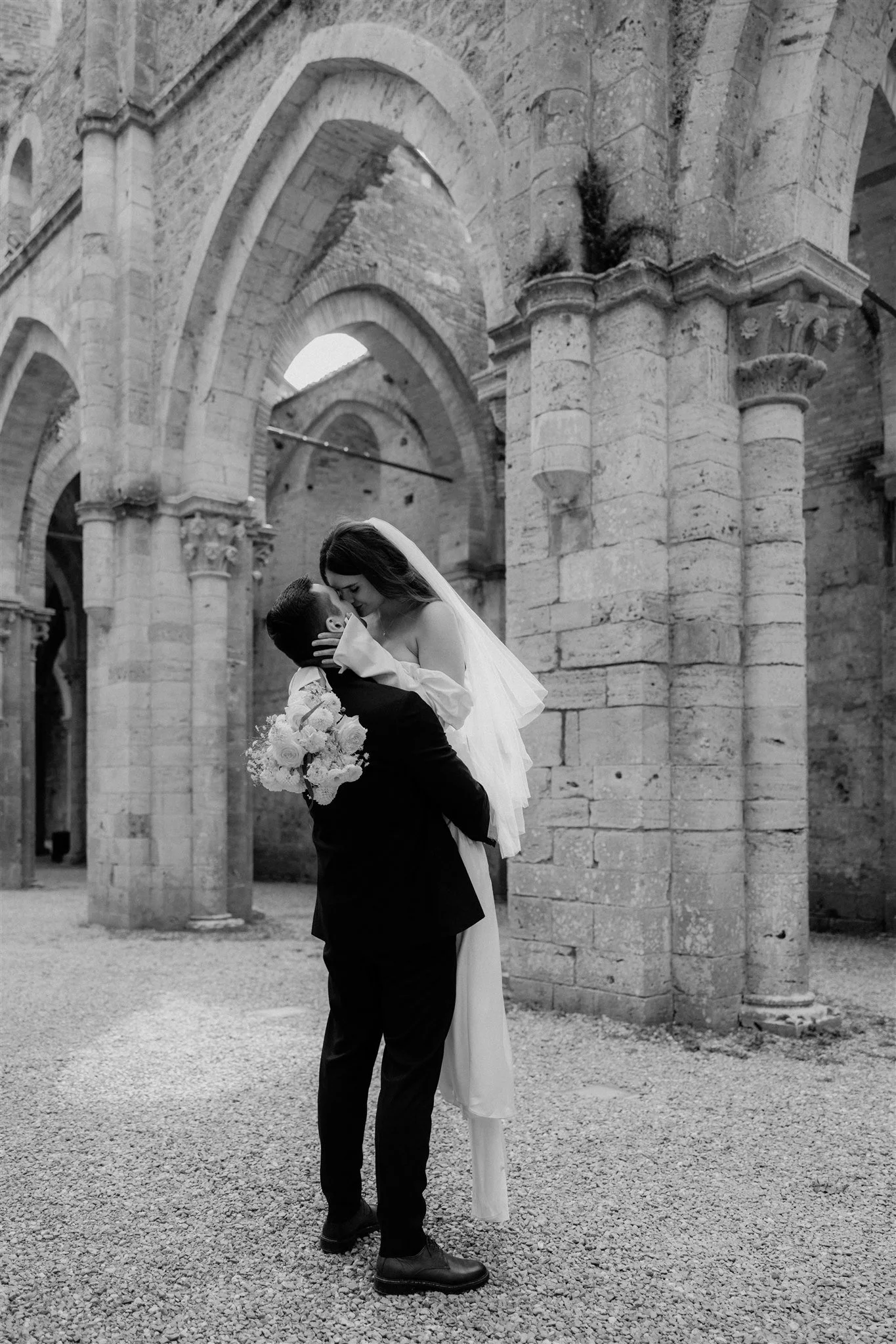 A couple dressed in wedding attire, embracing and about to kiss, in front of ancient stone arches and columns, with one person holding a bouquet of flowers.