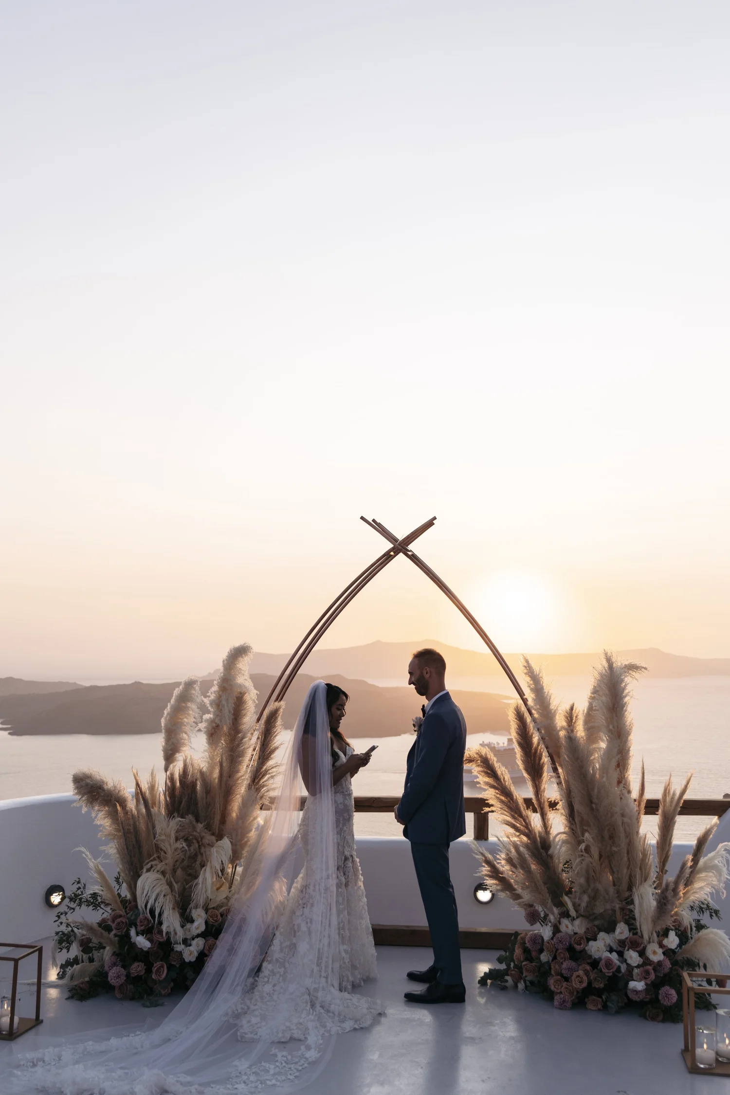 A couple getting married outdoors during sunset, standing under an archway decorated with pampas grass and flowers, with a scenic view of a body of water and mountains in the background.