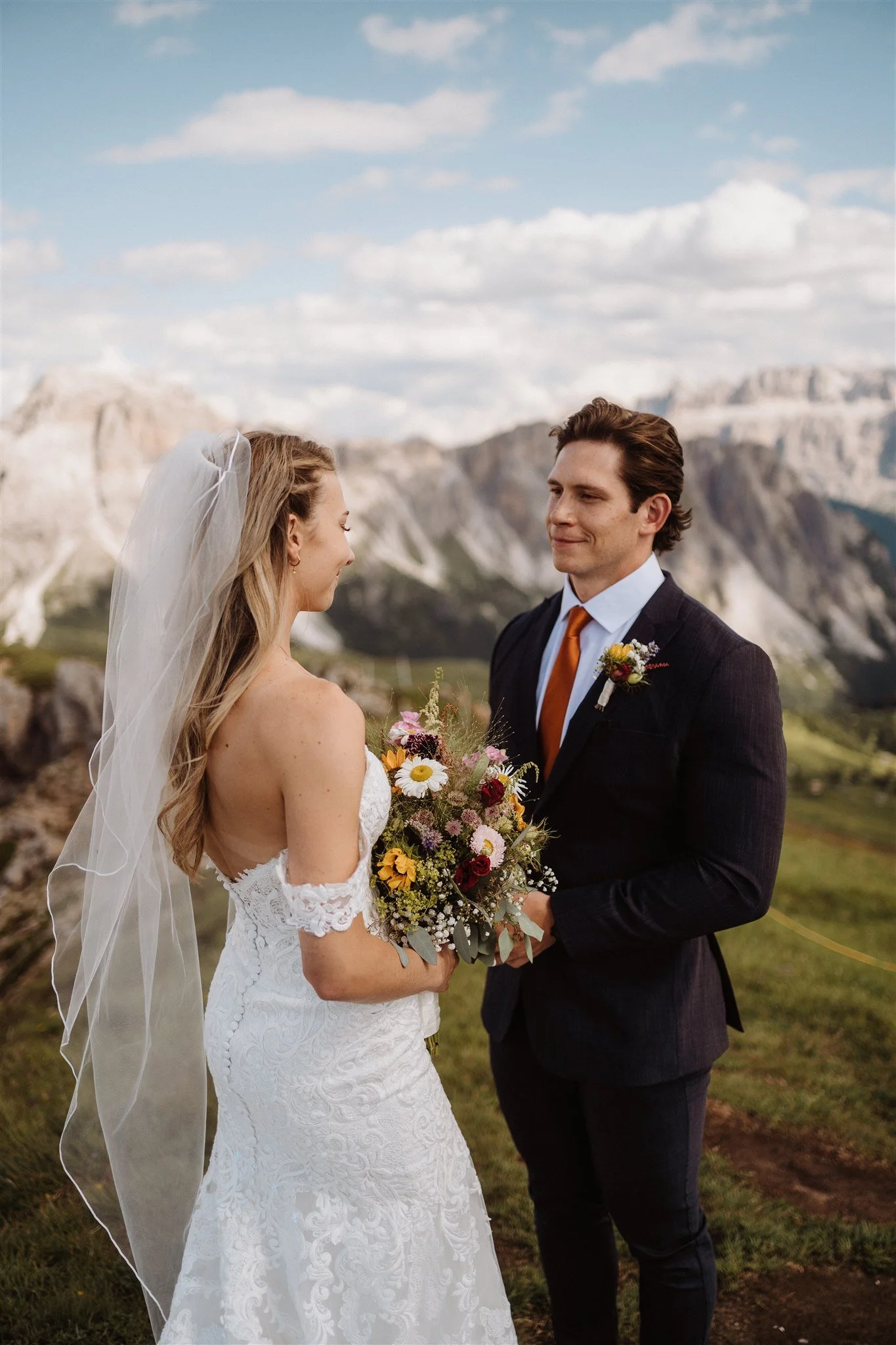 A bride and groom stand outdoors during a wedding ceremony, facing each other with mountain scenery and a partly cloudy sky in the background.