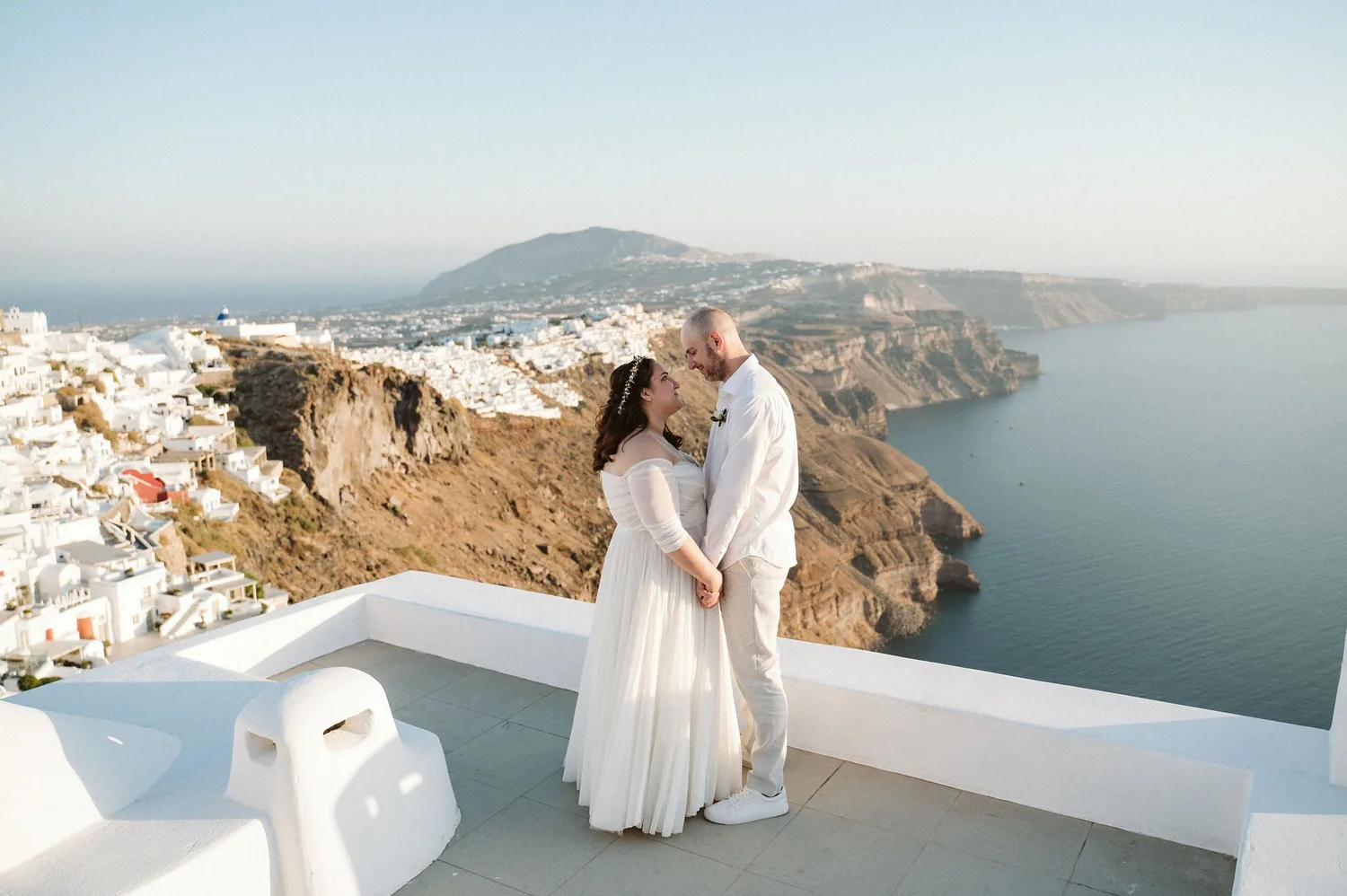 A couple in wedding attire standing on a white terrace overlooking the sea and a hillside town with white buildings, with a mountain in the background.