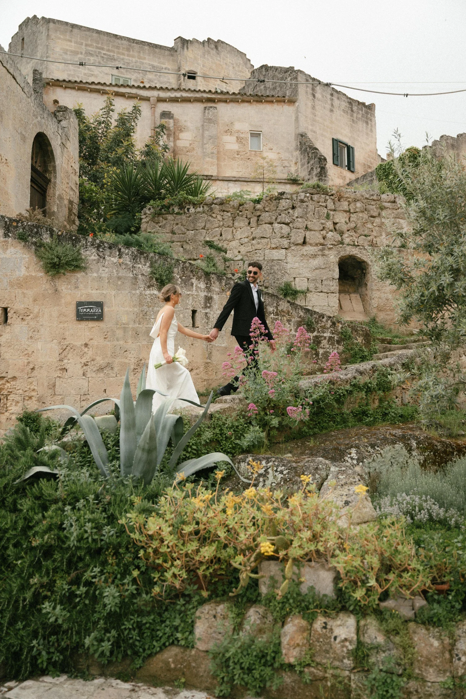 A couple holding hands and walking through a garden with colorful flowers and green plants, with old stone buildings in the background.