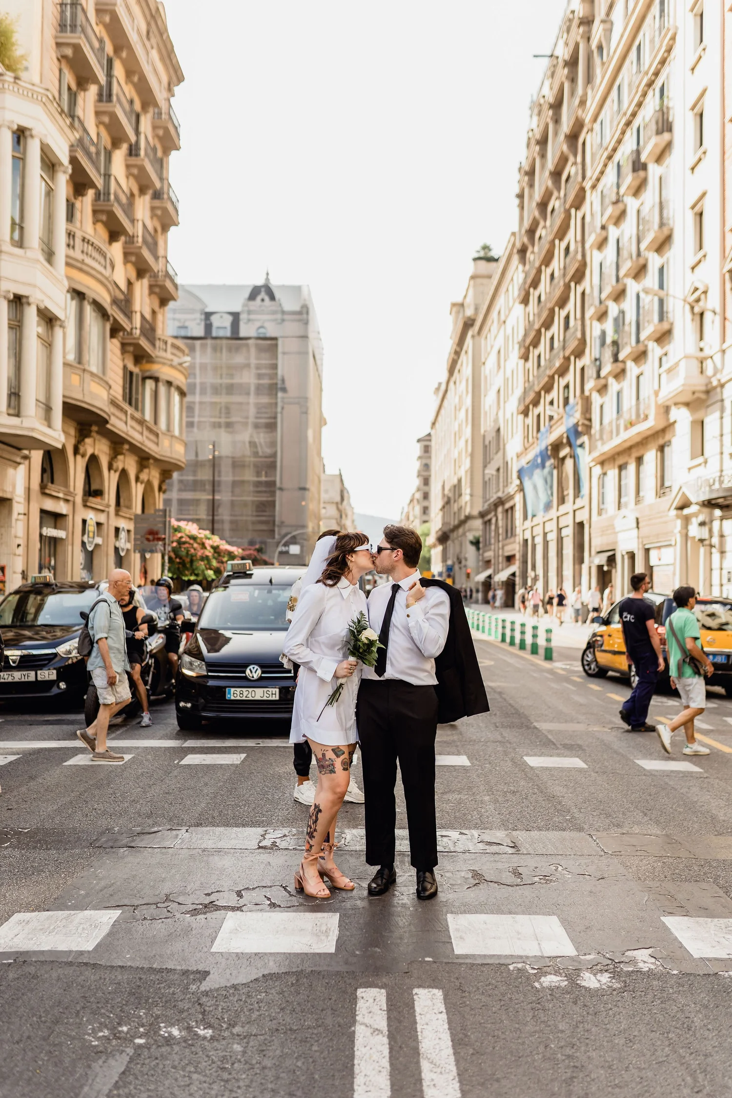 A bride and groom sharing a kiss on a city street during daylight, surrounded by pedestrians and cars, with historic buildings in the background.
