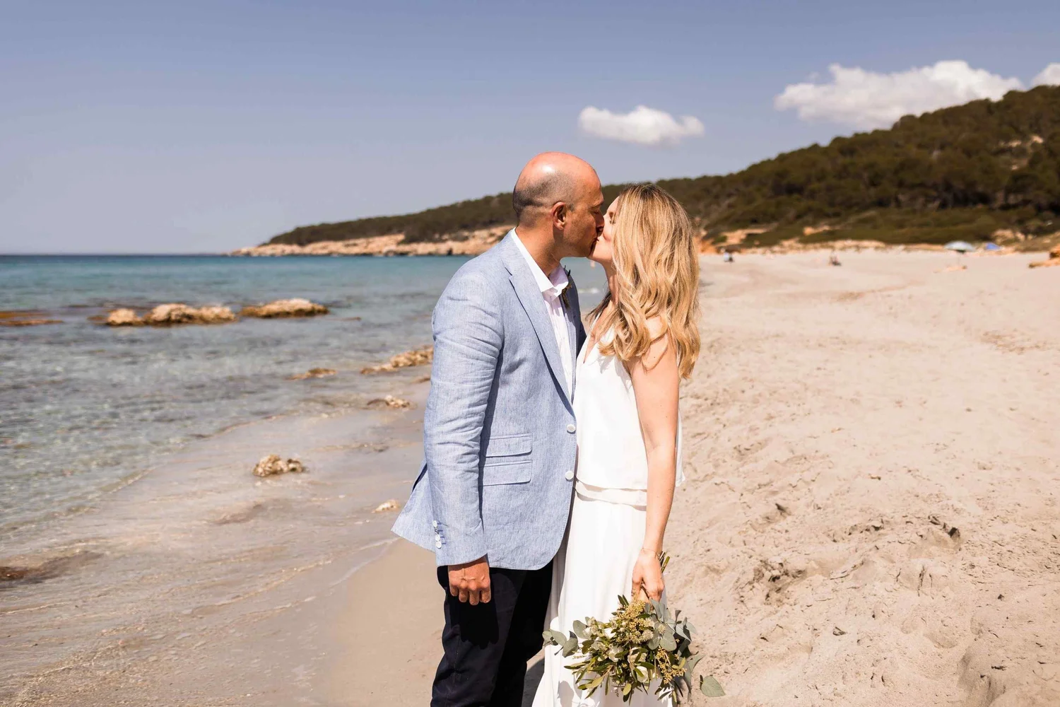 A couple sharing a kiss on the beach, with the man in a light gray blazer and the woman in a white dress holding a bouquet of greenery, ocean and shoreline in the background.