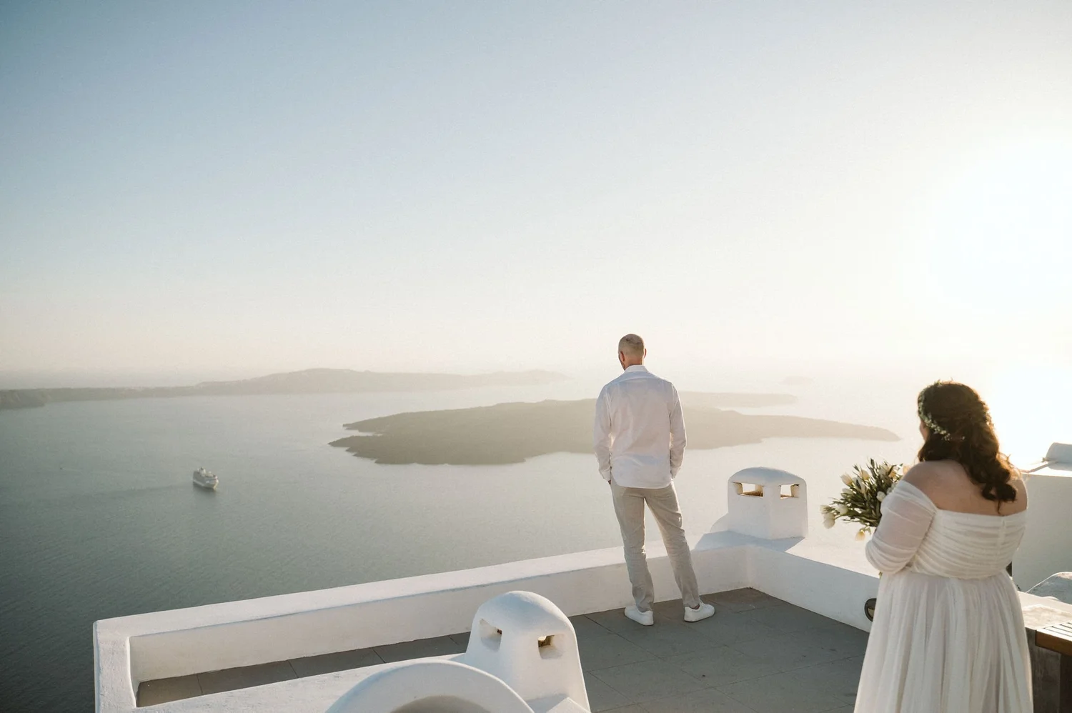 A man in a white shirt and pants standing on a balcony overlooking the ocean during sunset, with a woman in a white dress sitting nearby holding flowers.