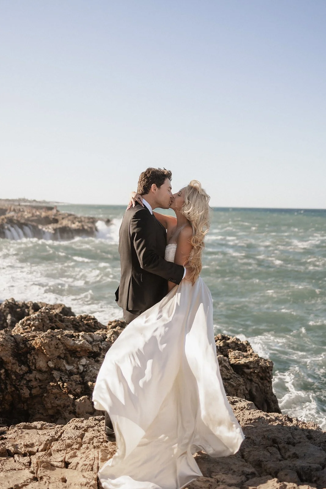 A bride and groom kissing on rocks by the ocean, with the bride in a white dress and the groom in a black suit.