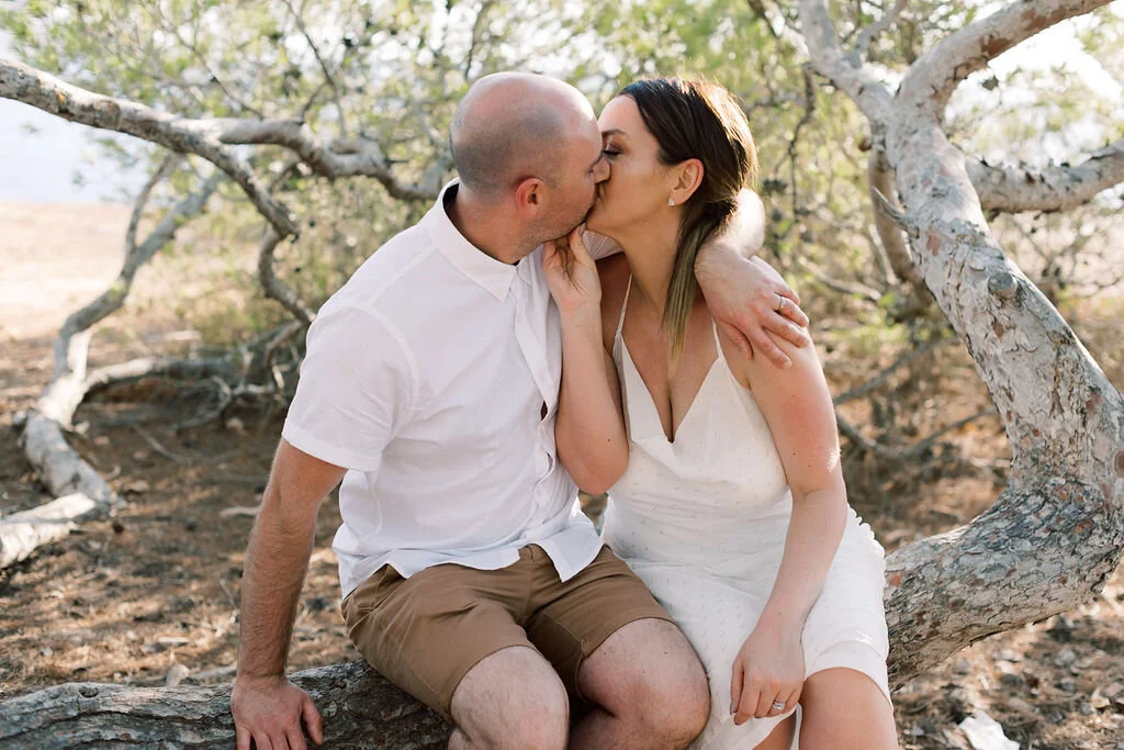 A couple sits on a tree branch outdoors, kissing, with the woman wearing a white dress and the man in a white shirt and tan shorts.