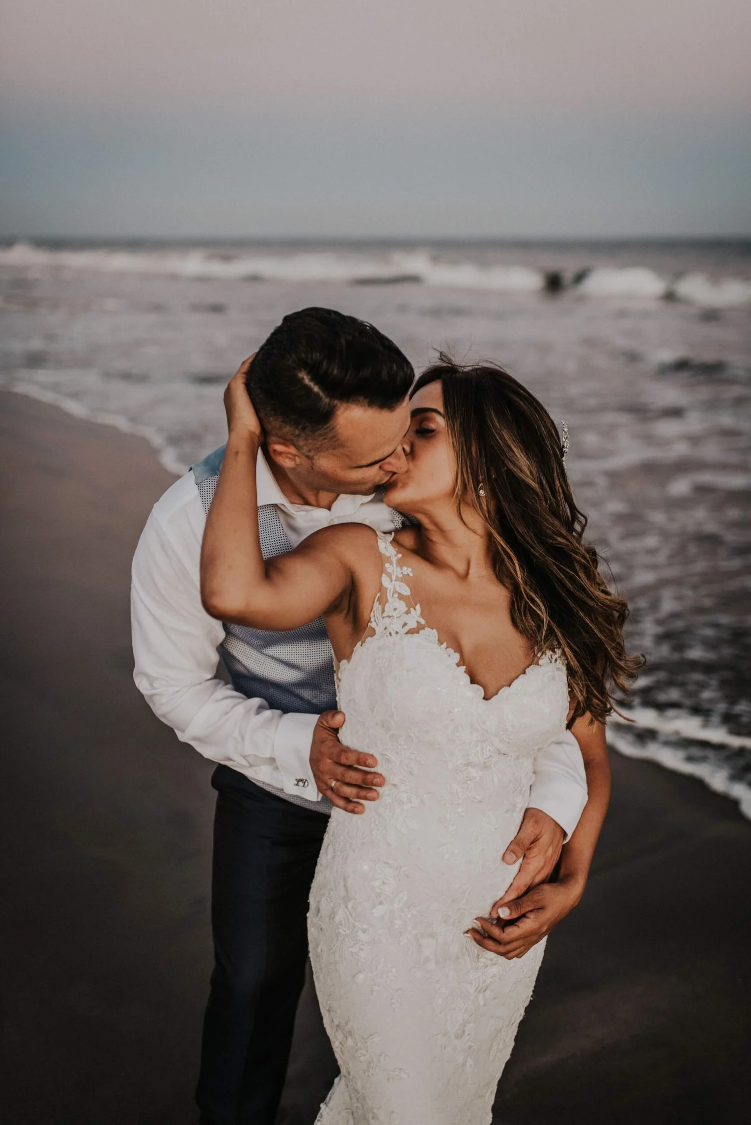 A bride and groom kissing on a beach during sunset. The groom wears a white shirt with suspenders, and the bride wears a white lace wedding gown. The ocean waves are in the background.