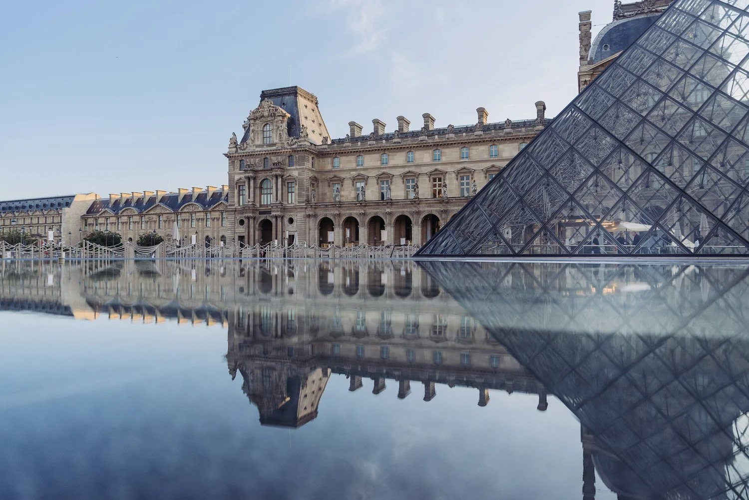 Historic building with ornate architecture and a glass pyramid structure in front, reflected in a water surface.