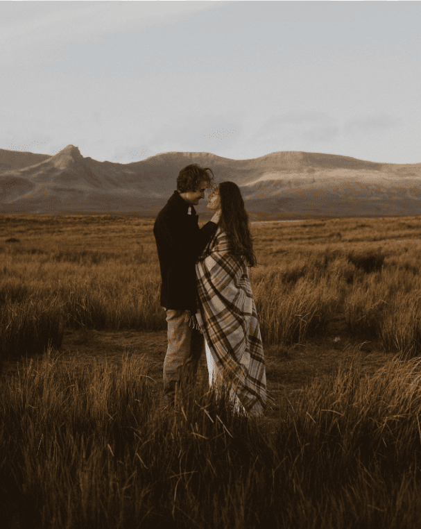 A couple stands close together in a grassy field with mountains in the background, sharing an intimate moment.