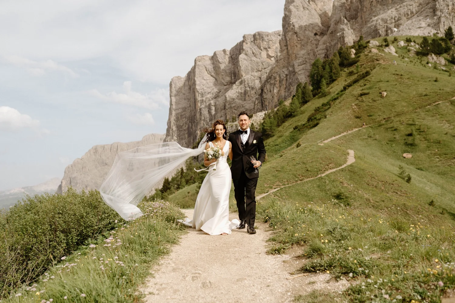 A bride and groom walking along a mountain trail in a scenic outdoor setting with green hills and rocky cliffs.