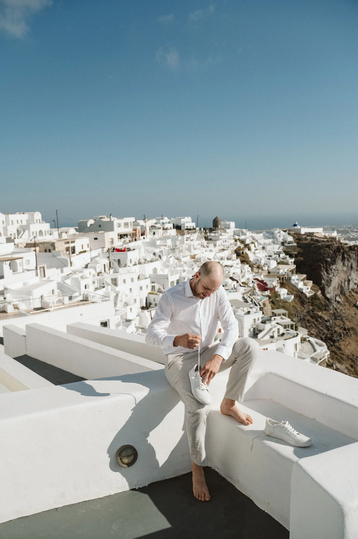 A man is sitting barefoot on a white ledge on a sunny day, tying his shoe with a background of white buildings and blue sky in a coastal town.