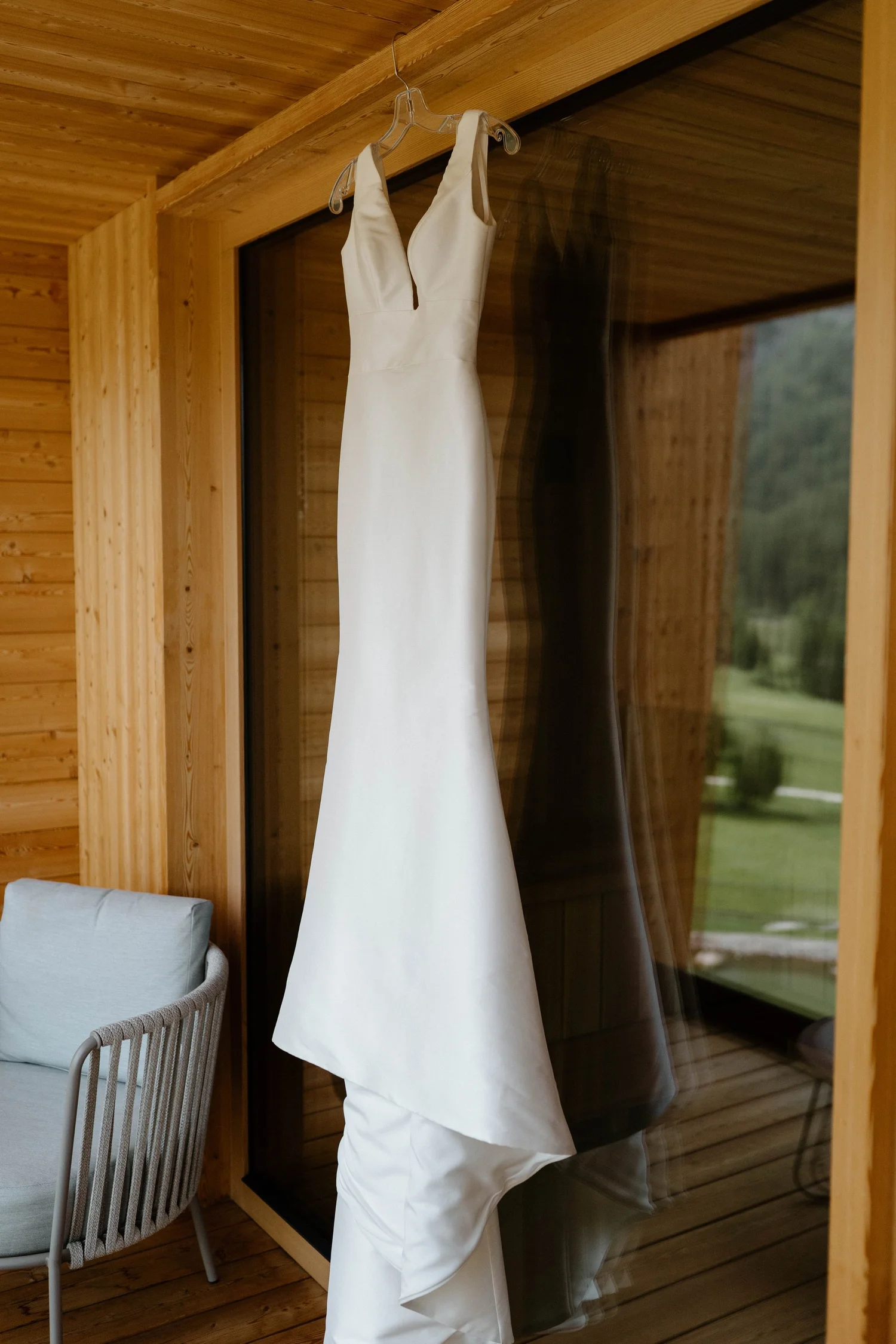 A white wedding dress hanging on a clear hanger inside a modern wooden house, with a reflection visible on a glass door.