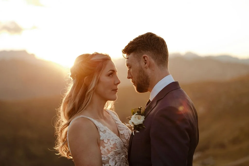 A bride and groom face each other outdoors during sunset, with the sunlight behind them.