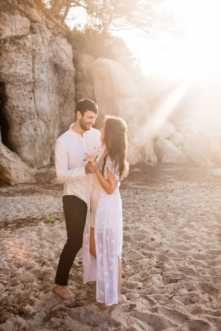 A young couple dancing and smiling on a sandy beach with large rocks in the background, illuminated by warm sunlight.