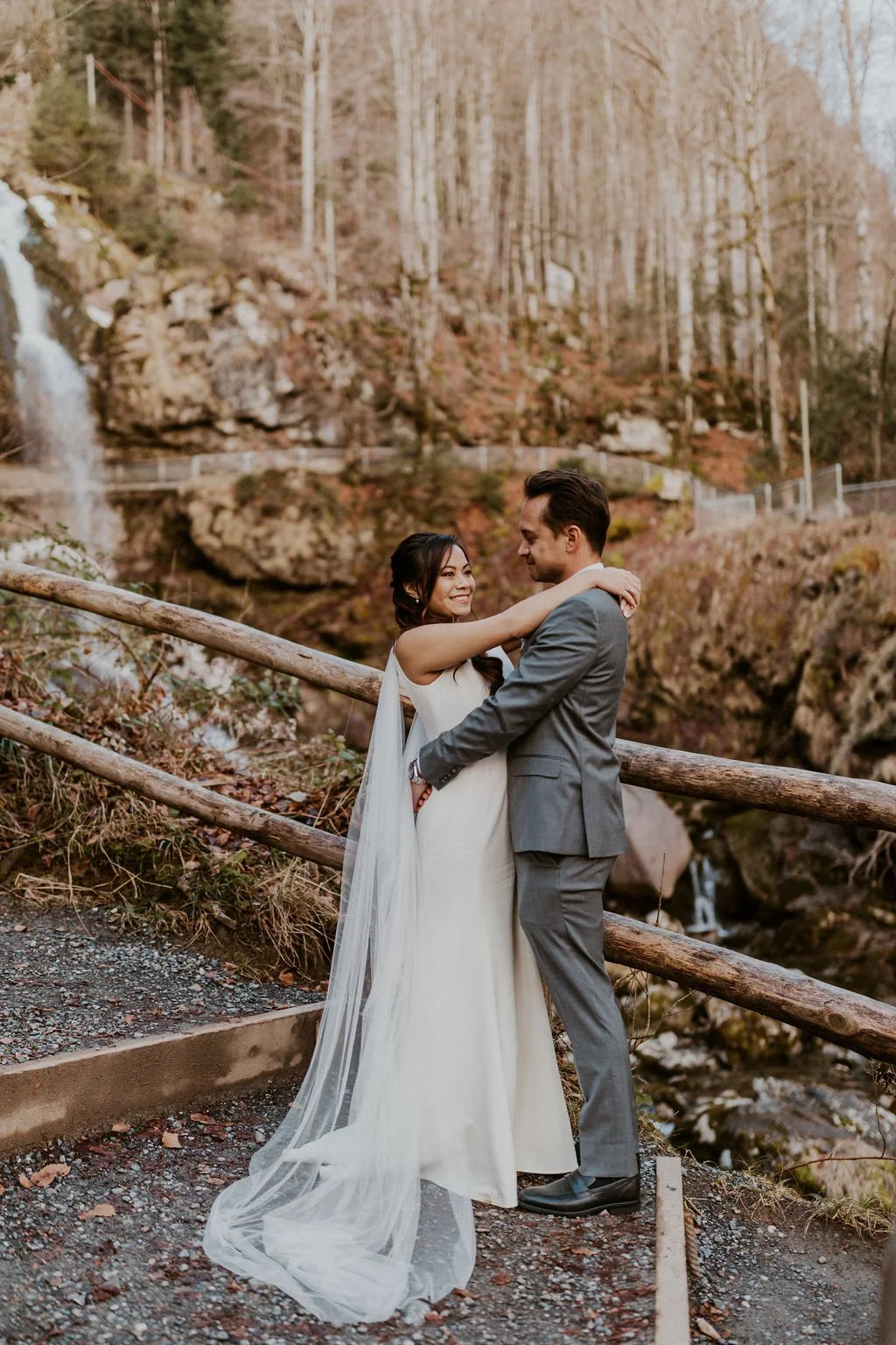 A bride and groom embrace on a rocky trail with a wooden railing, in front of a natural landscape with a waterfall, rocks, and leafless trees in the background.
