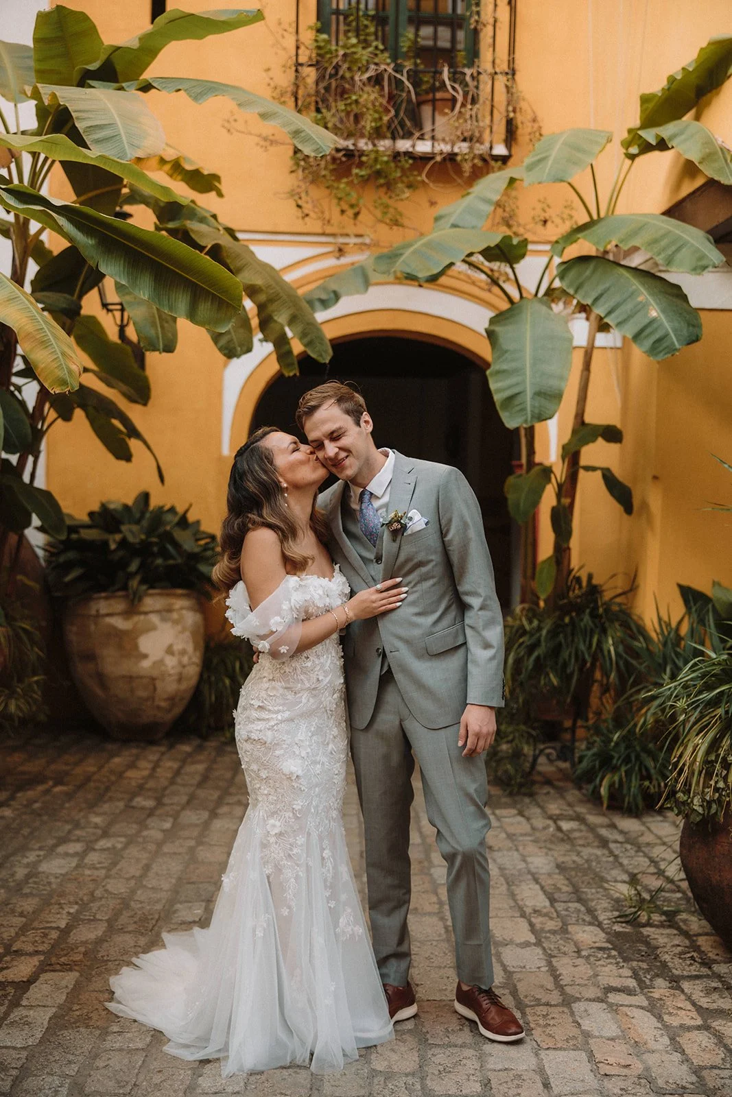 A bride and groom sharing a loving moment in an outdoor courtyard with large potted plants and lush greenery.
