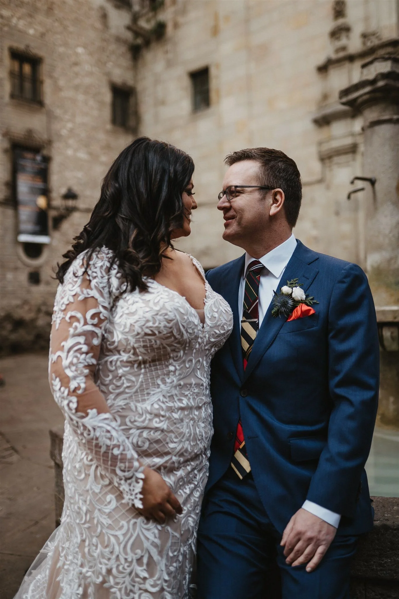 A couple dressed in wedding attire standing close and smiling at each other, outdoors with an old stone building in the background.